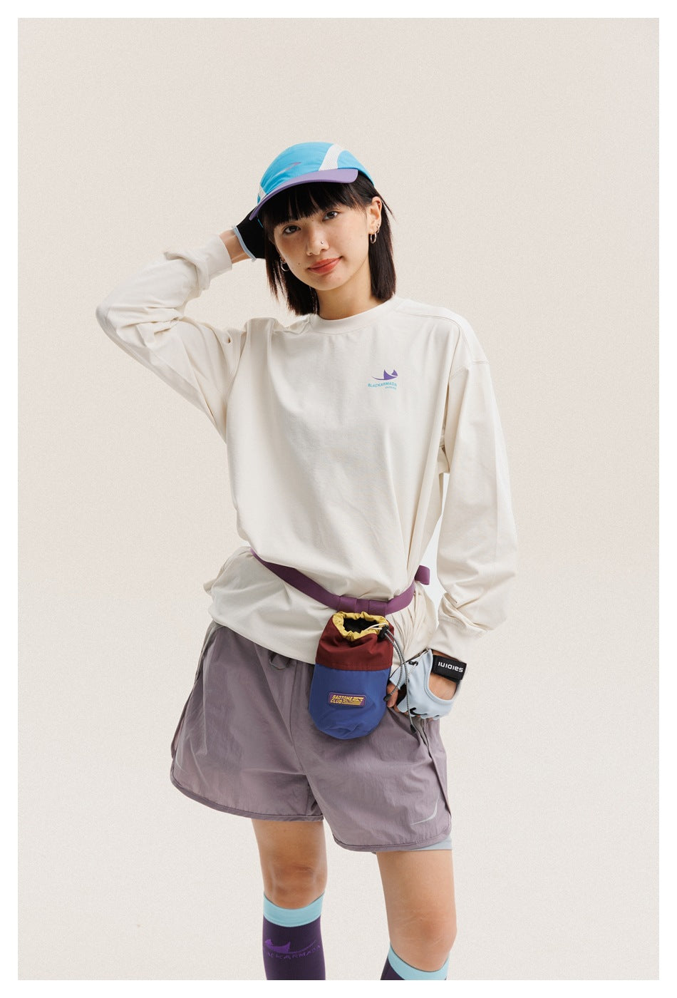 Model in a cream long-sleeve shirt and gray-mauve shorts with a blue cap, gloves, and waist pouch.