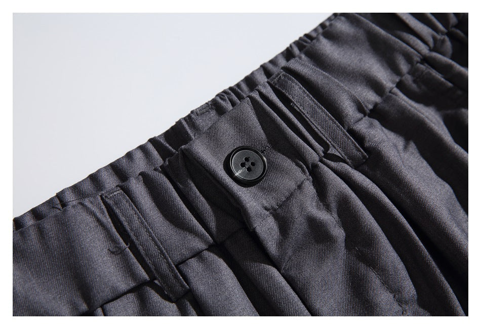 Close-up of charcoal trousers with a buttoned waistband tab, belt loops and front pleats.