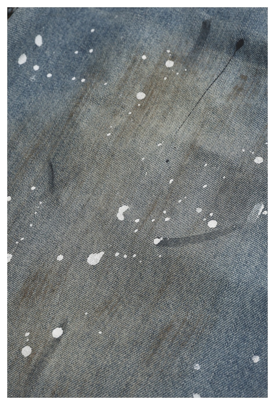 Close-up of faded blue denim with scattered white paint splatters and dark distress marks.