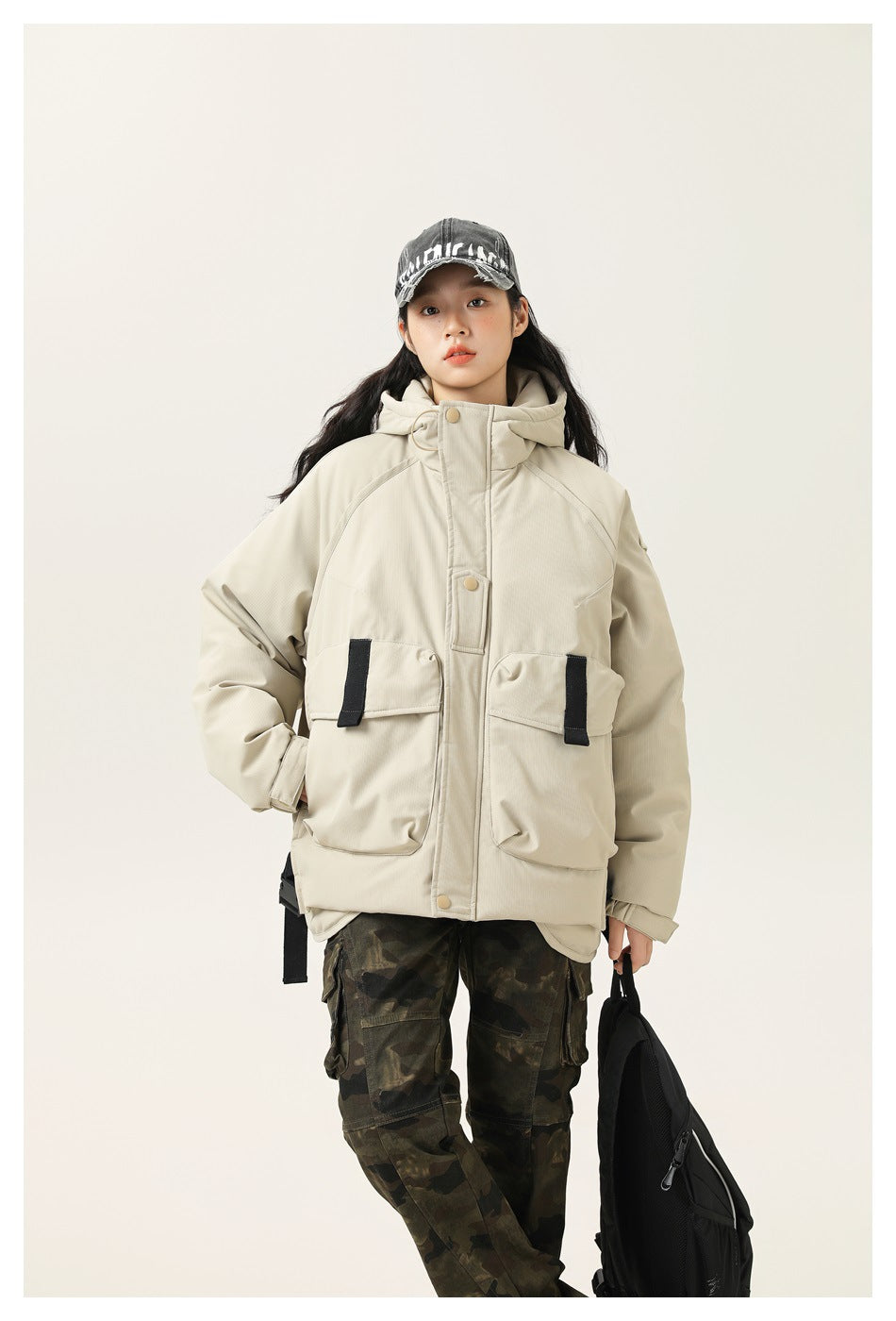 Model wearing a zipped light cream hooded jacket with large flap pockets and black accents, styled with camouflage pants.