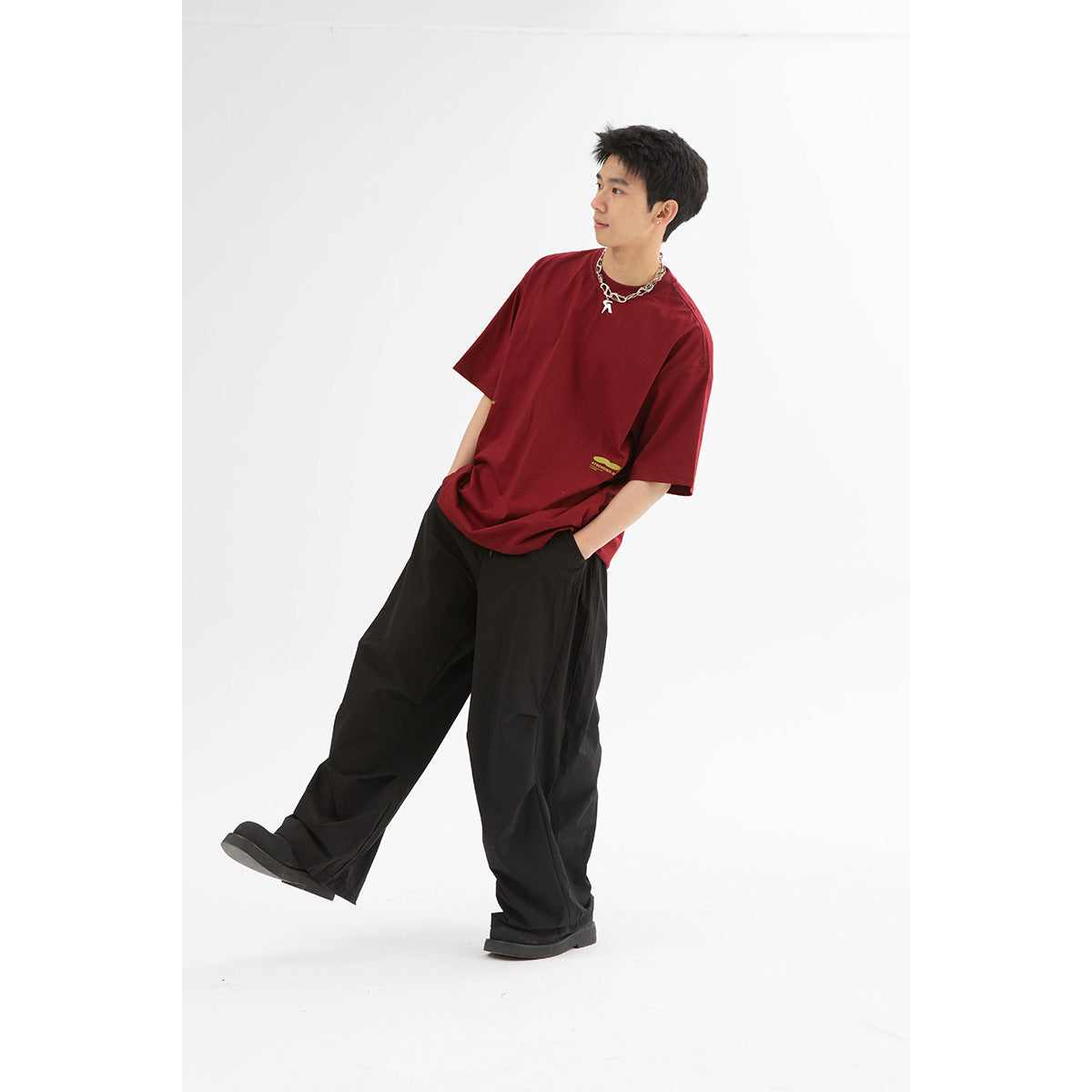 Model wearing an oversized burgundy T-shirt with a small yellow chest graphic and loose black trousers.