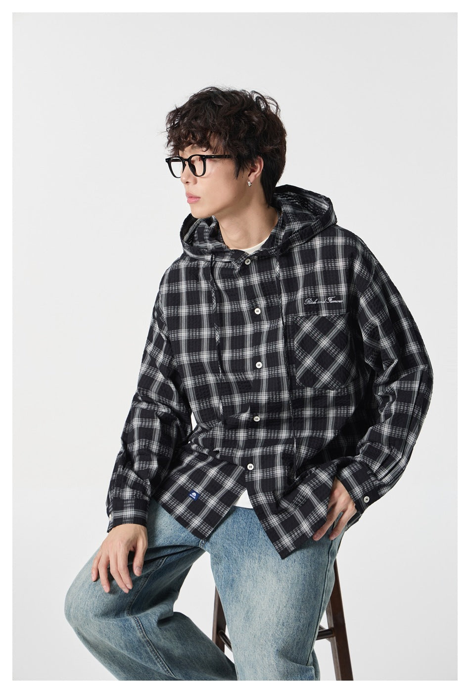 Seated model wearing a black-and-white plaid hooded button-up shirt with a chest pocket and blue jeans.