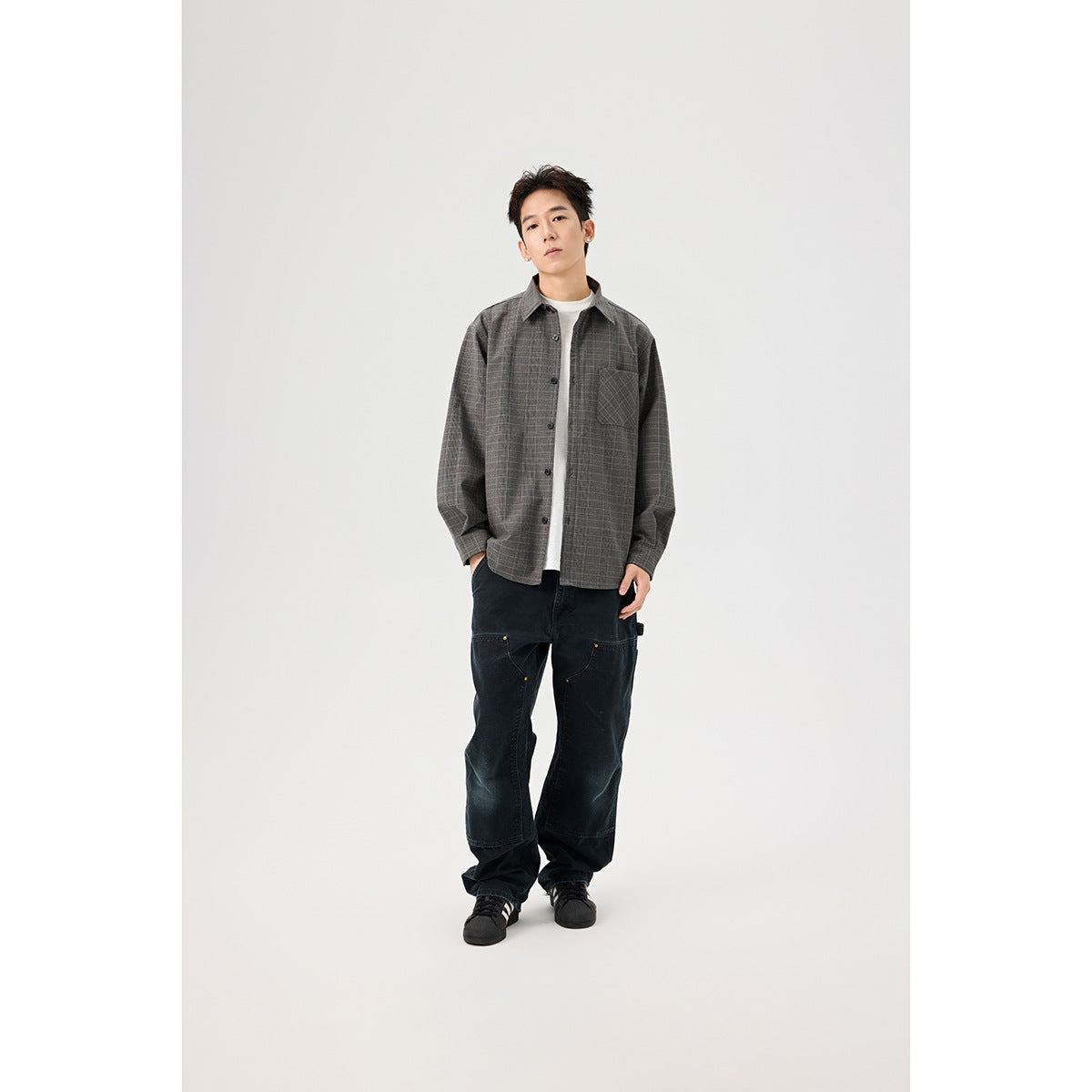 Model wearing a gray plaid button-front overshirt with a chest pocket over a white tee and loose black pants.