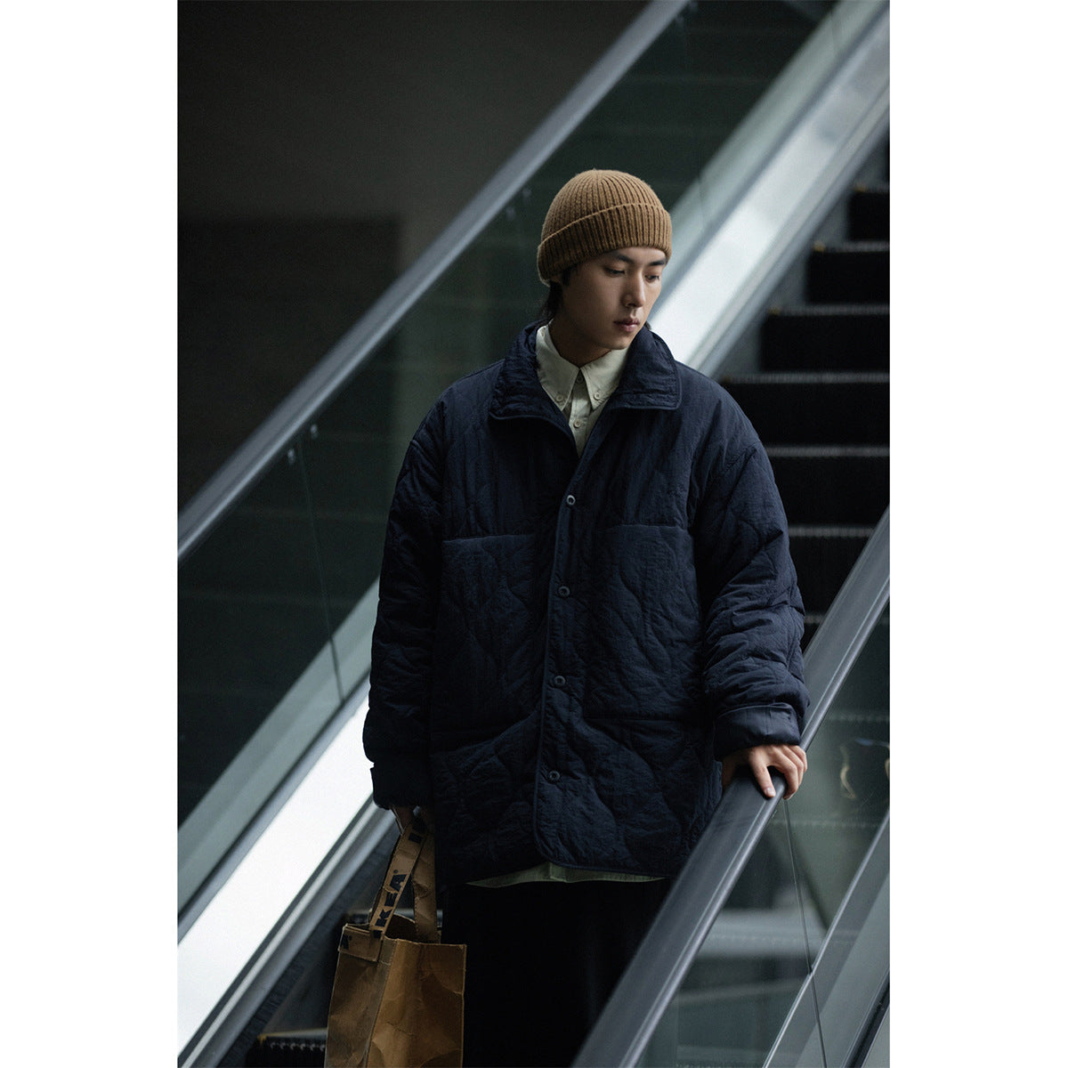 Model wearing a dark navy quilted button-front coat with a wide collar and a brown knit beanie on an escalator.