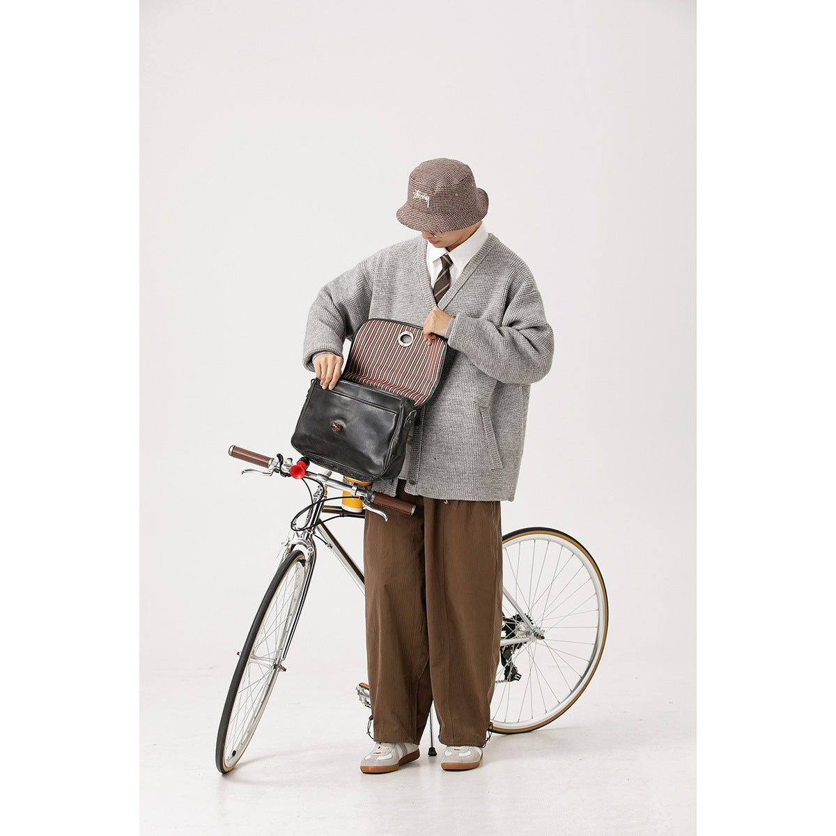 Model wearing a light gray textured V-neck cardigan with brown trousers, opening a black bag beside a bicycle.