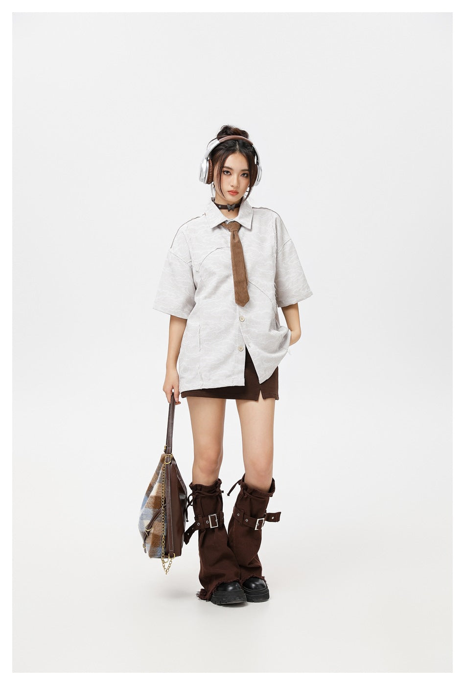 Front view of a model wearing an oversized white patterned short-sleeve shirt, brown tie, and dark mini skirt.