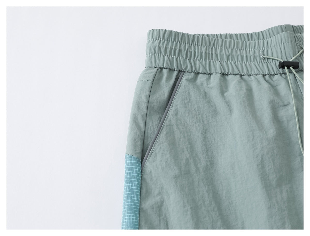 Close-up of sage green pants showing the elastic waist, angled front pocket, and blue grid-patterned side panel.
