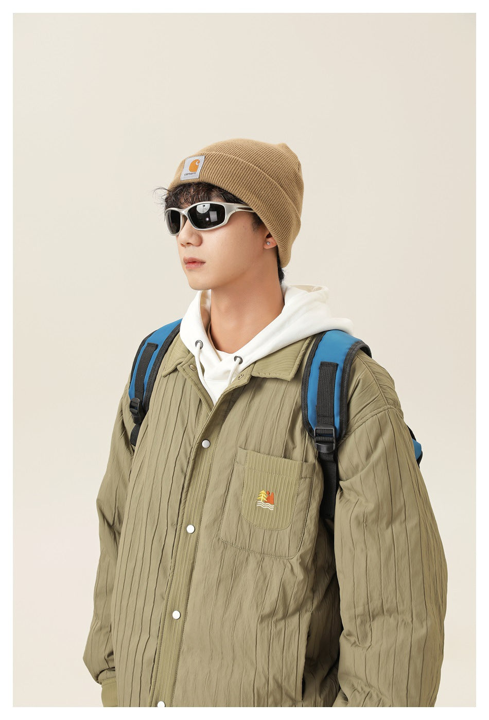 Model wearing an olive quilted snap-front jacket over a white hoodie, with a chest pocket and embroidered graphic.