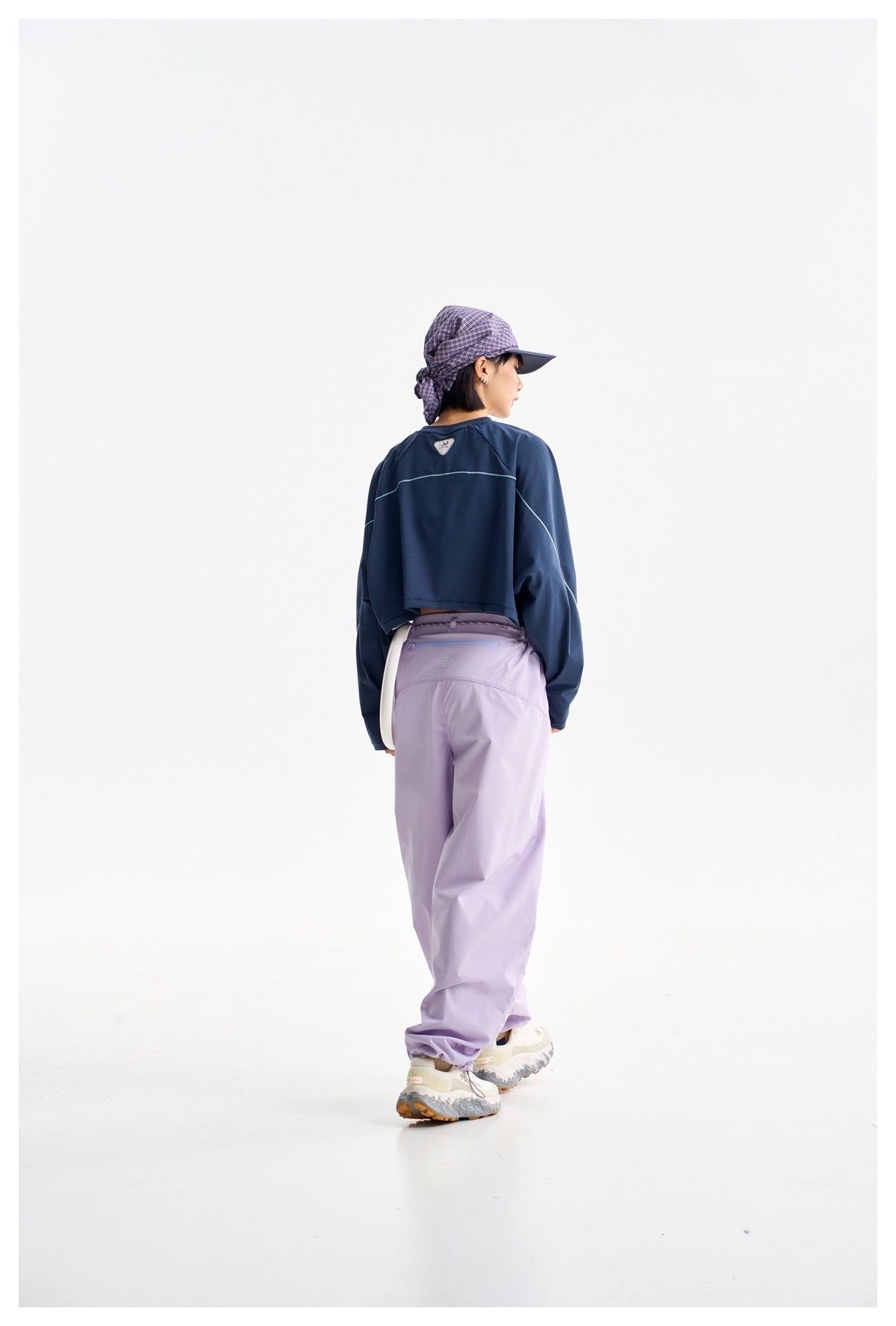 Rear view of a model wearing a navy cropped top and lilac pants with a plaid cap.