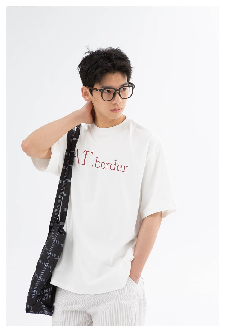 Model wearing a white oversized T-shirt with burgundy chest lettering, white pants, and a black plaid shoulder bag.