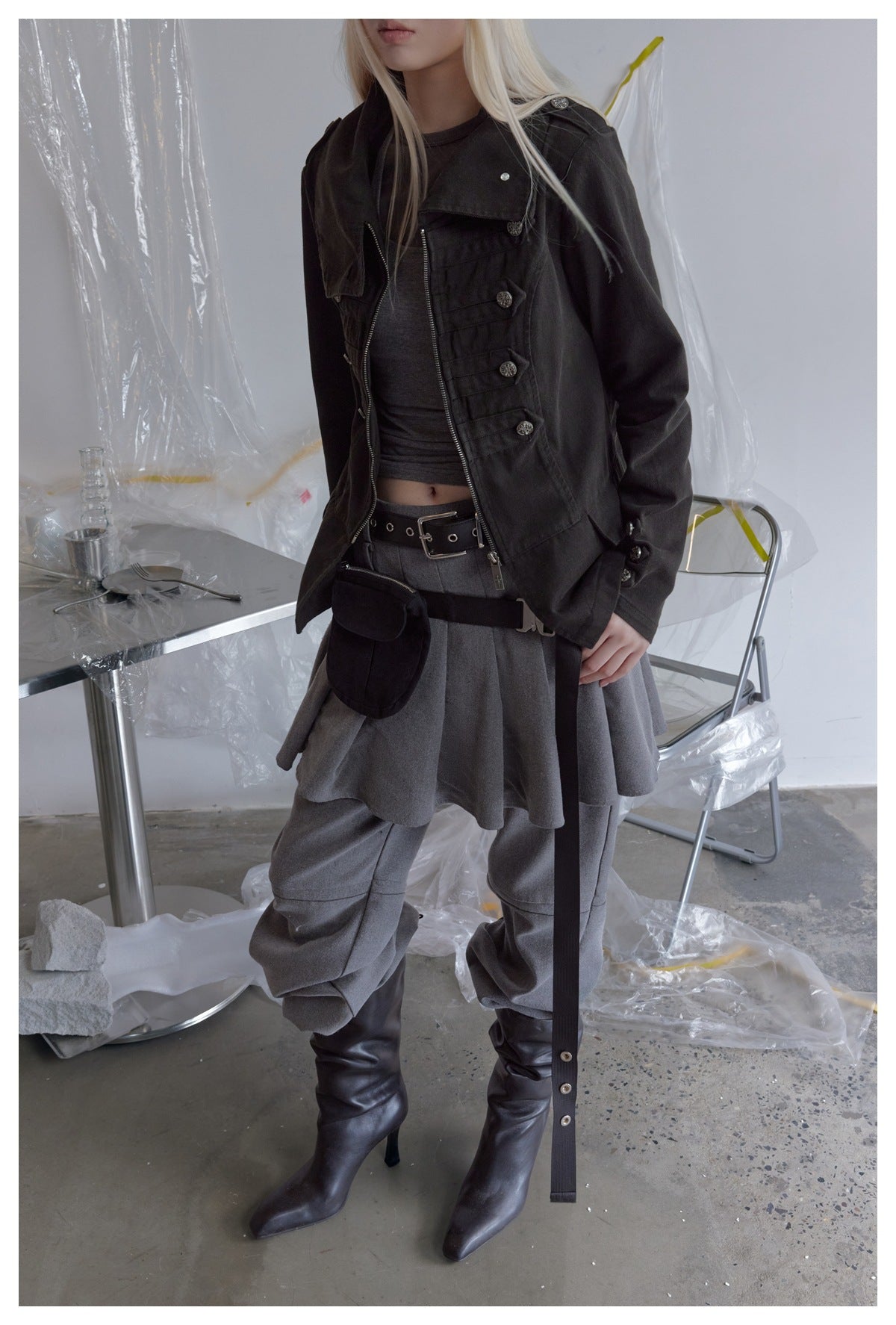 Cropped view of a model in the black buttoned jacket, gray tank top, gray pleated skirt, pouch, belt, and boots