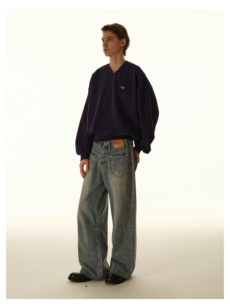 Model wearing an oversized dark V-neck sweatshirt with a small chest emblem and loose gray-blue jeans.