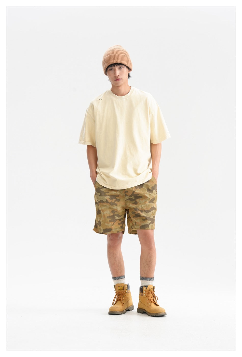 Man facing forward in a cream oversized T-shirt and tan camouflage shorts against a white studio background.