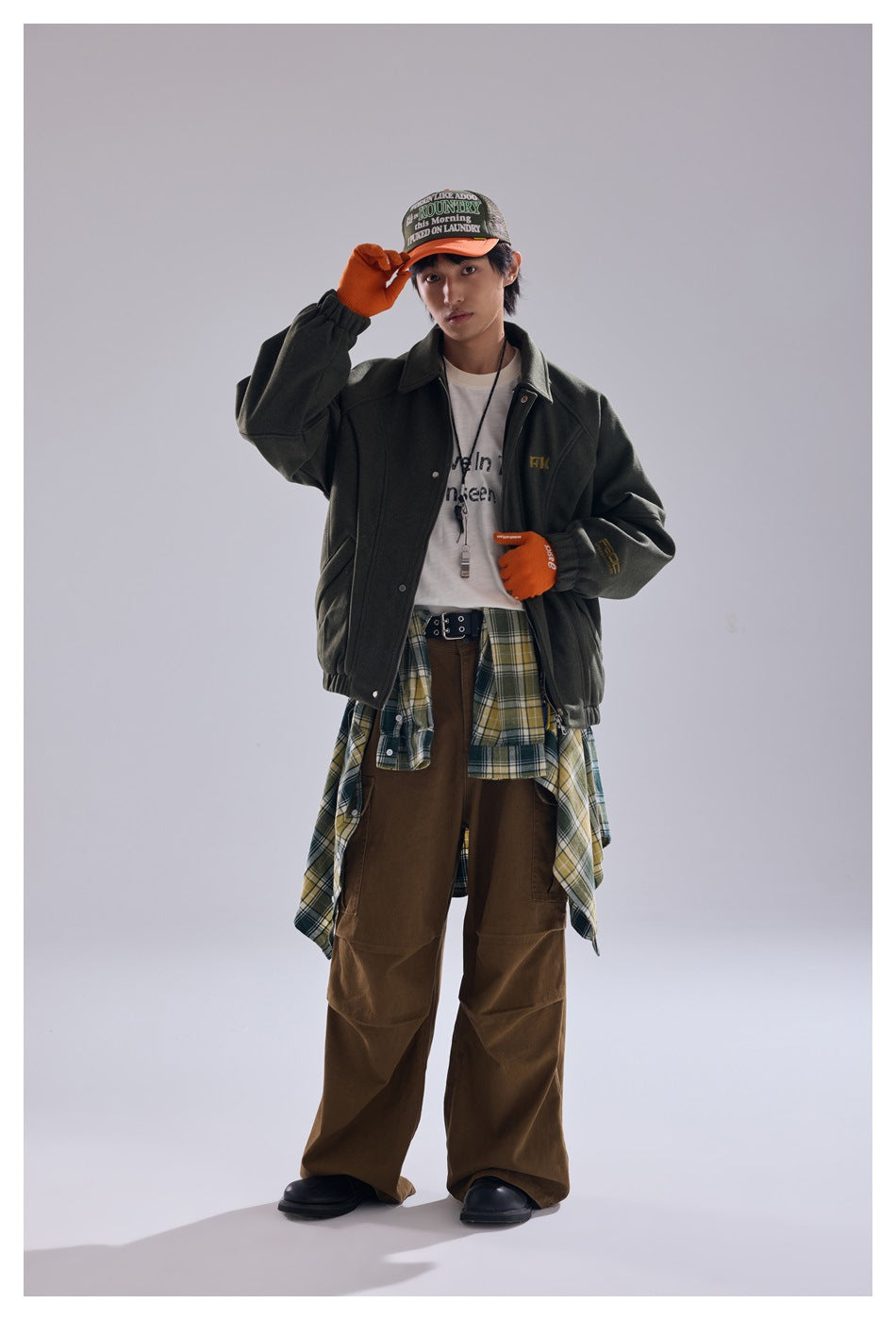 Model wearing an olive green jacket over a white tee with a plaid shirt tied at the waist and brown wide-leg pants