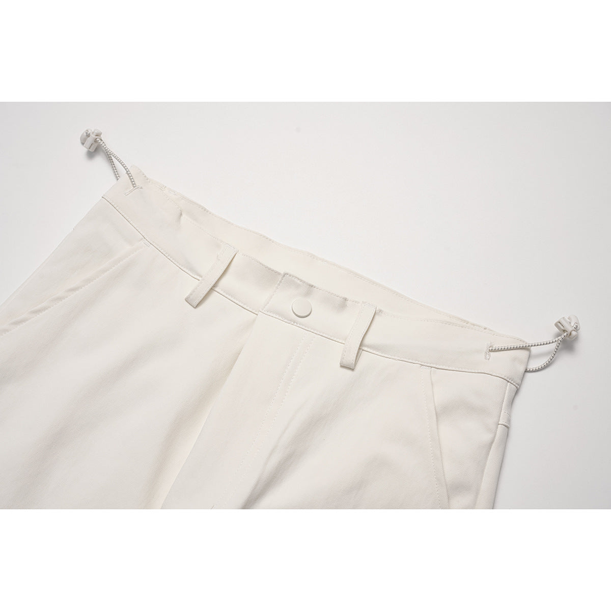 Close-up of cream pants waistband with button, belt loops, and adjustable side cords.