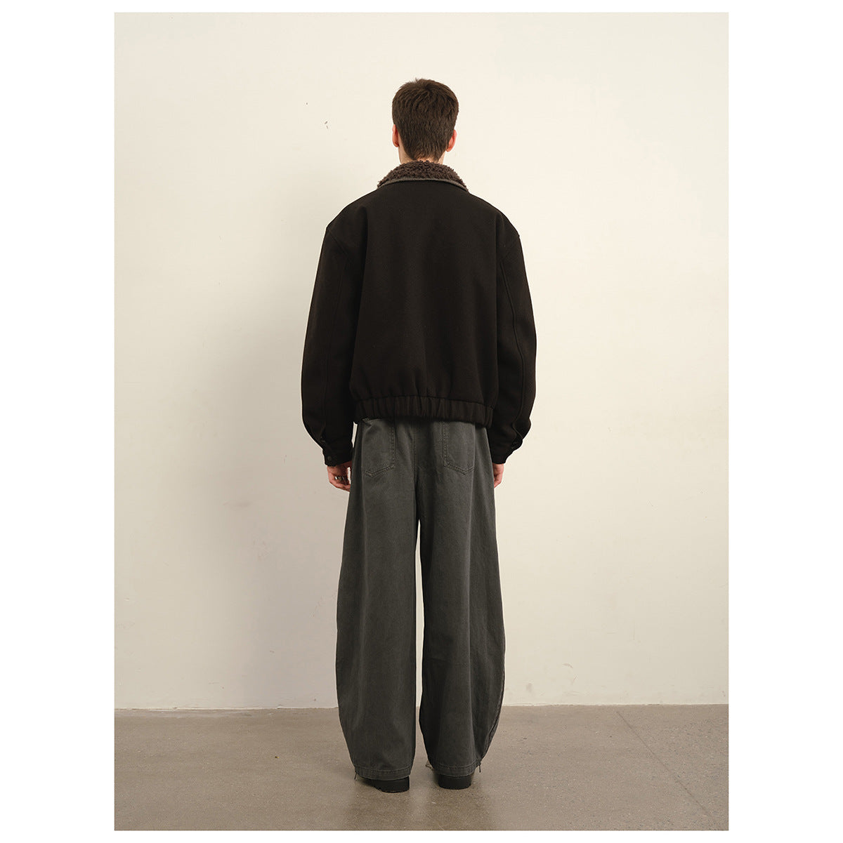 Back view of a model wearing a black jacket with a brown textured collar and wide gray jeans.