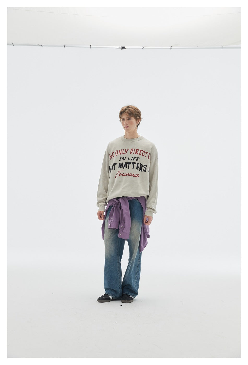 Model standing in a beige slogan knit sweater, faded blue jeans and a purple shirt tied at the waist