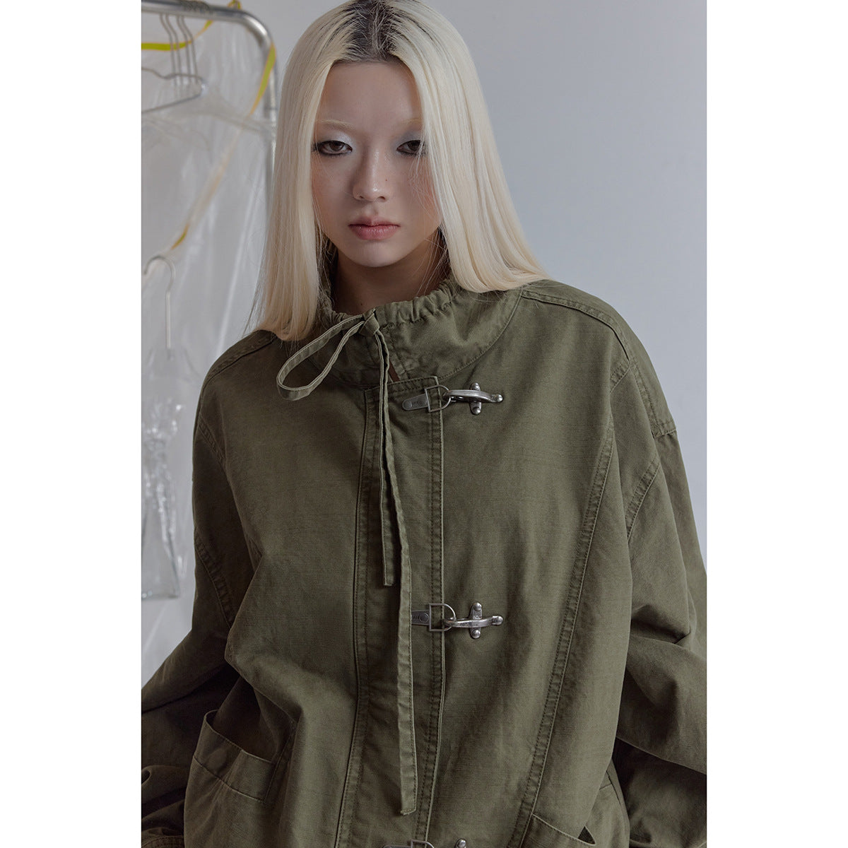 Model in a close view wearing an olive jacket with a gathered drawstring collar and silver hook closures.