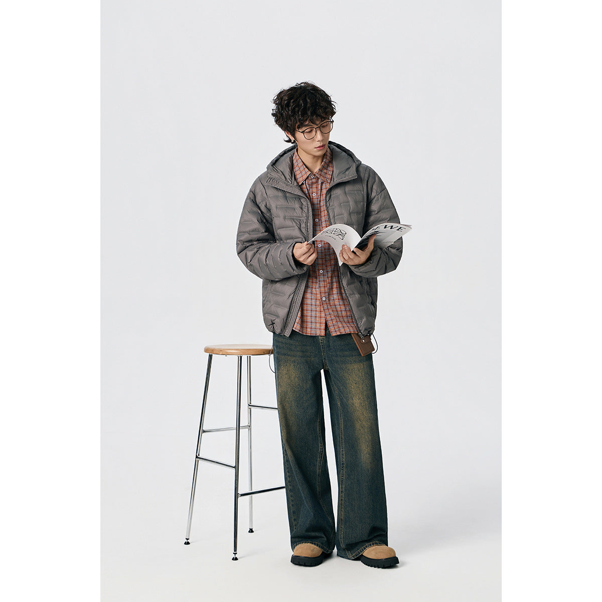 Model wearing a gray hooded quilted jacket over a plaid shirt, shown full length while reading a magazine.