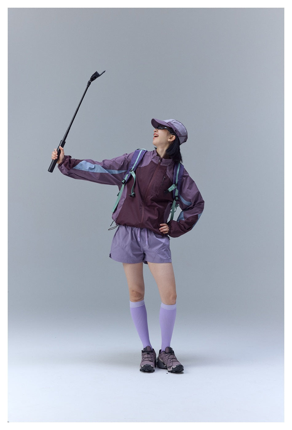 Model in a plum color-block zip jacket, lavender shorts and cap, posing with a selfie stick.