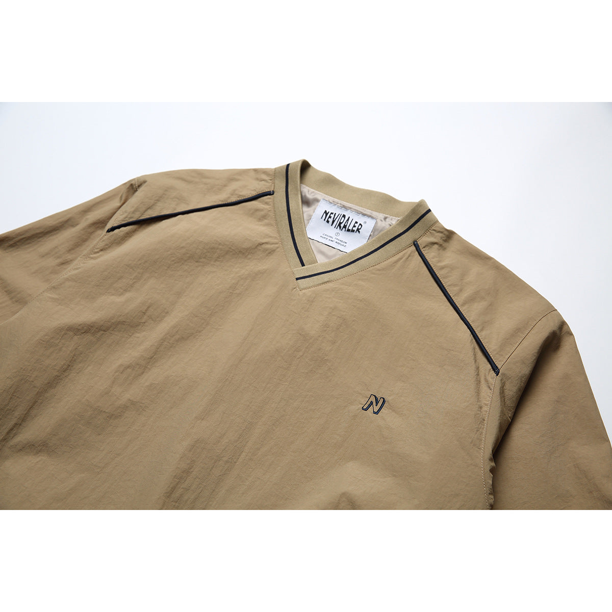 Close-up of a tan V-neck jacket front with dark piping, a small embroidered N, and a visible neck label.