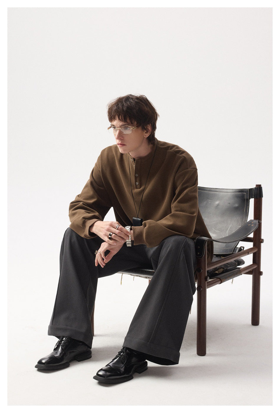 Seated full-length model wearing a brown henley top, charcoal wide-leg trousers, and black shoes.