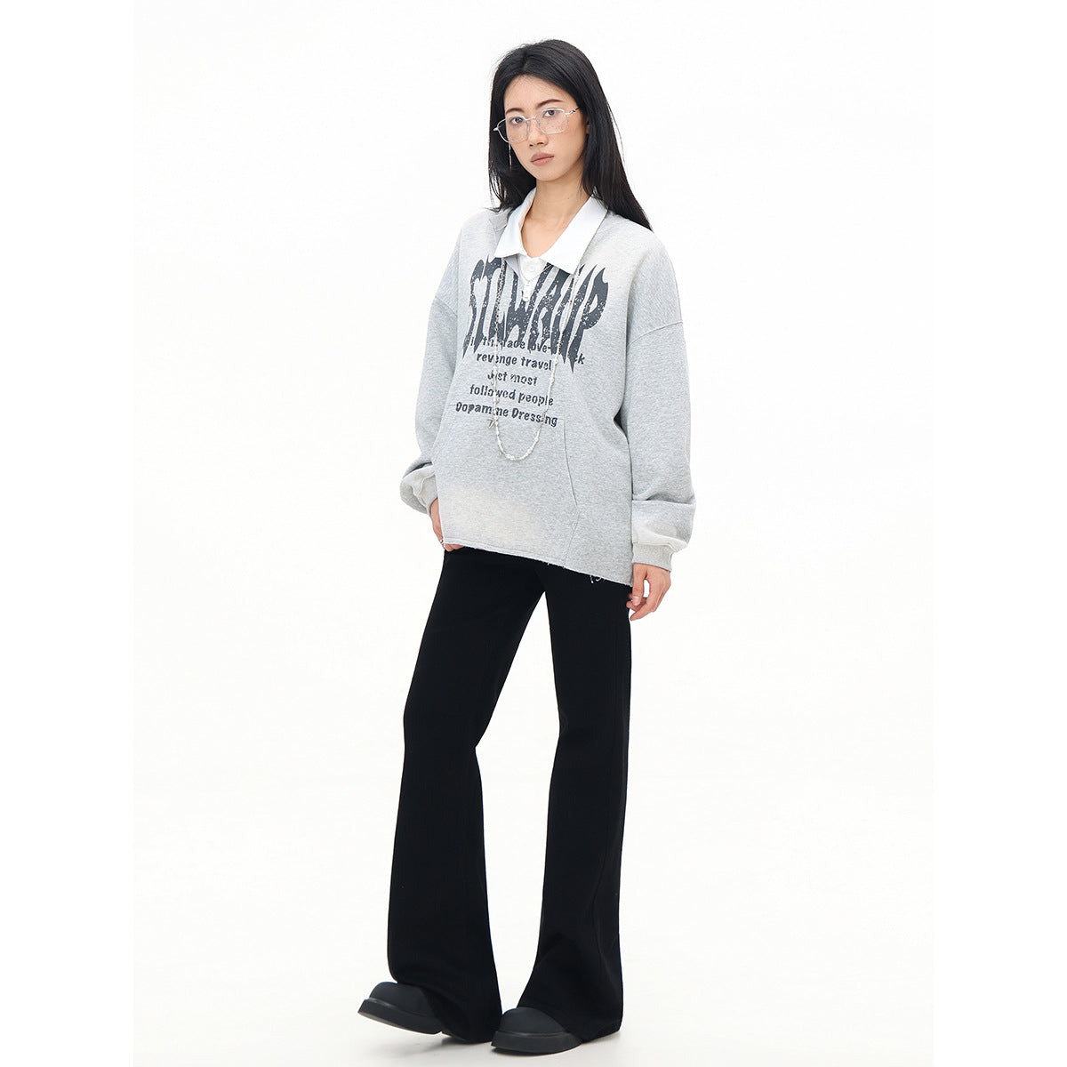 Model wearing a light gray graphic sweatshirt over a white collared shirt with black flared pants, shown full length.