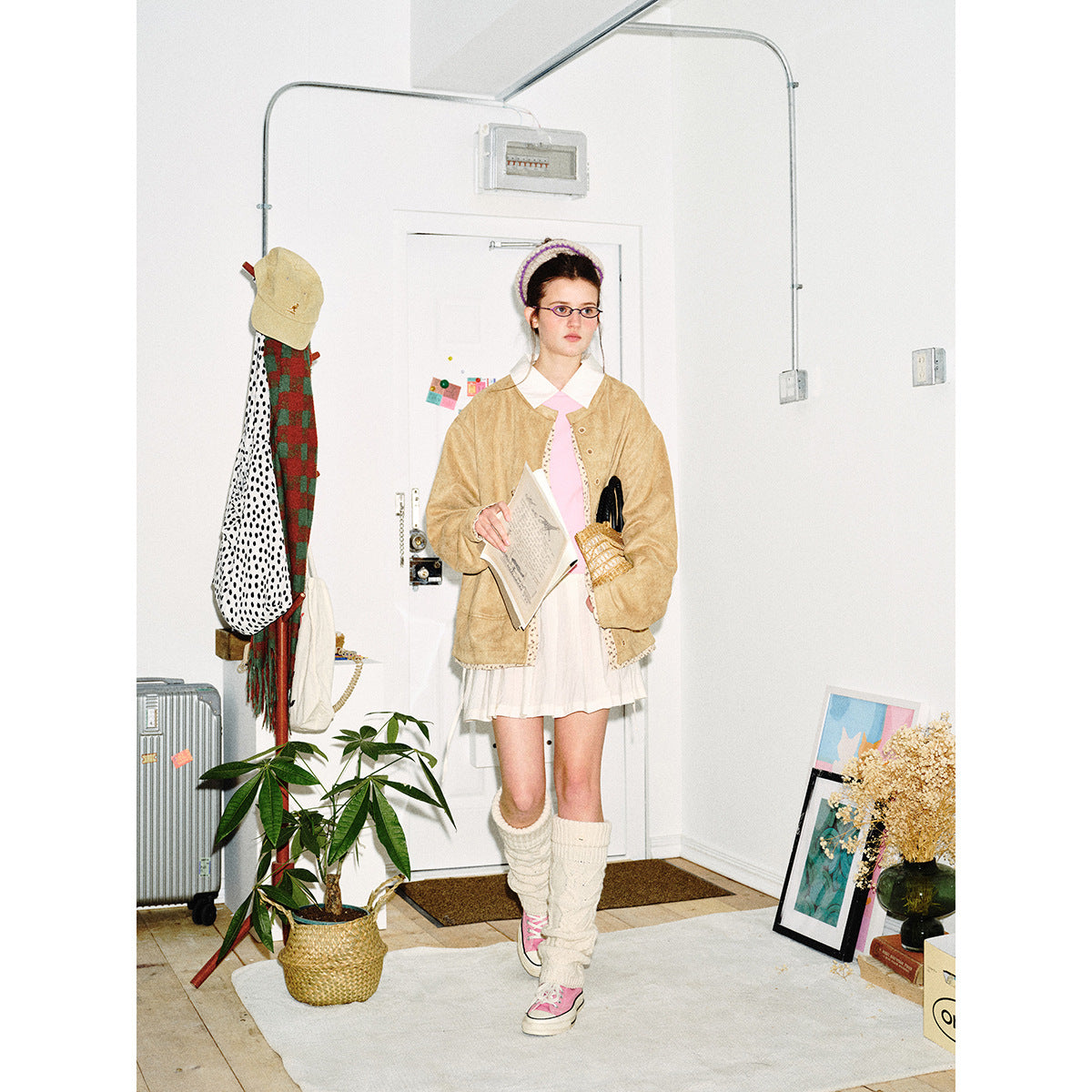 Model wearing a tan jacket with patterned trim over a white outfit and pink tie, facing forward in a room.