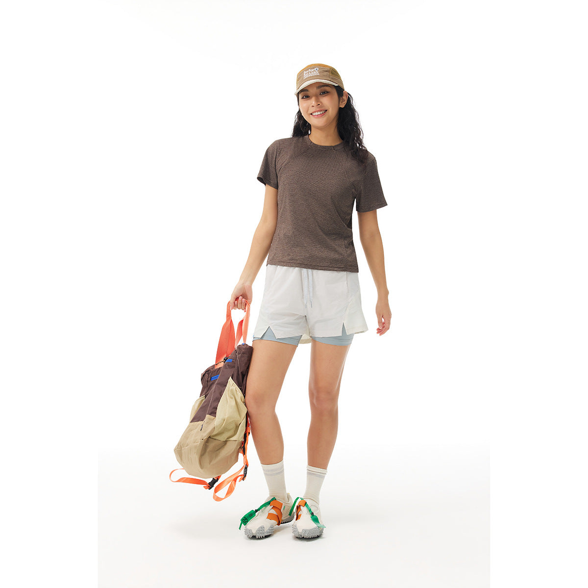 Model wearing a brown short-sleeve shirt and white layered athletic shorts, carrying a bag.