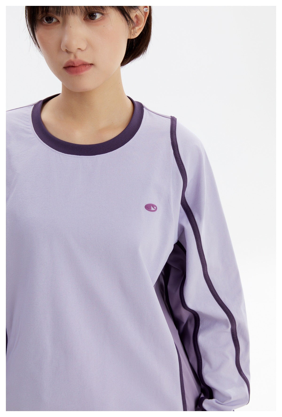 Close-up of a lavender top showing its dark purple crew neck, small chest emblem, and contrasting sleeve trim.