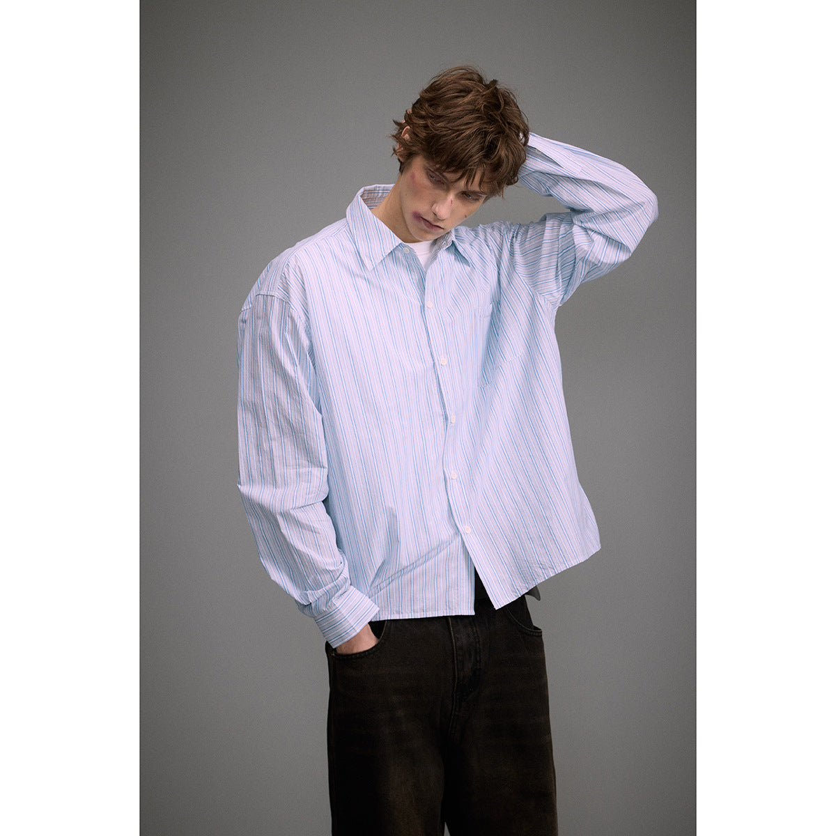 Model wearing a loose white button-up shirt with thin blue stripes, a chest pocket, and black jeans.