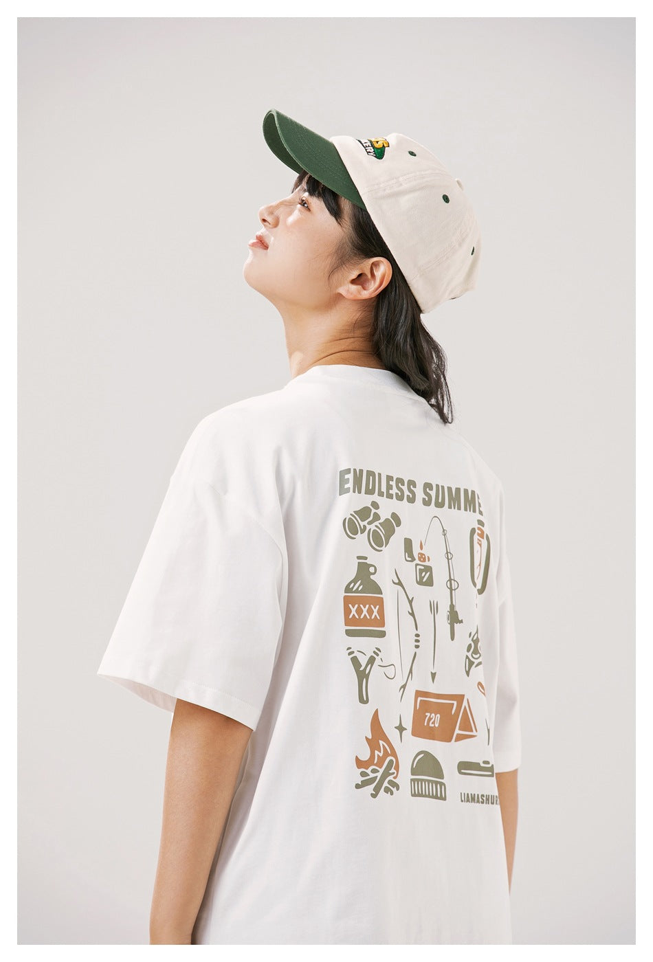 Close view of a white T-shirt's large camping-themed back graphic, worn with a cream cap with a green brim.