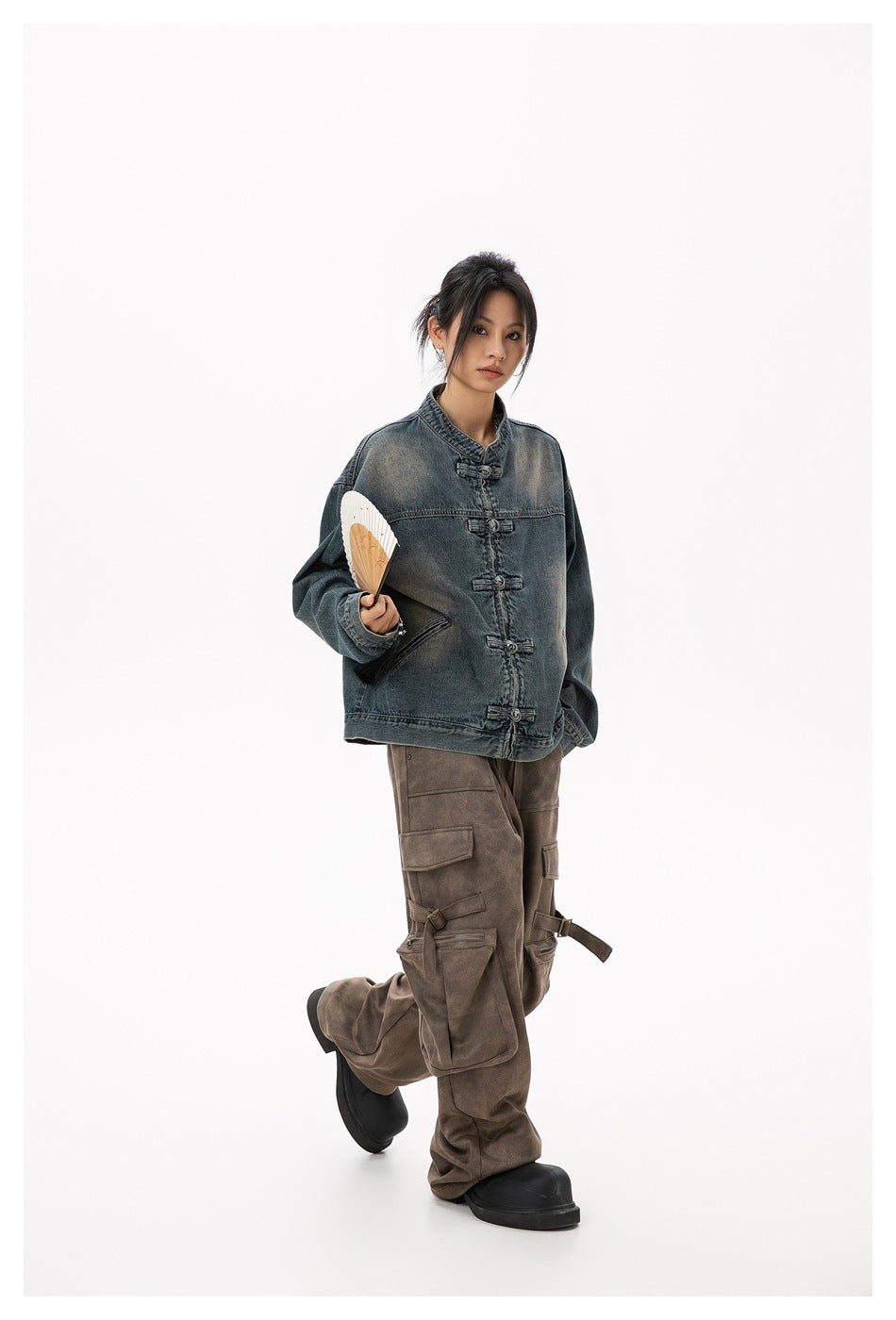Model wearing a faded blue-gray denim jacket with frog closures and brown cargo pants, holding a folding fan.