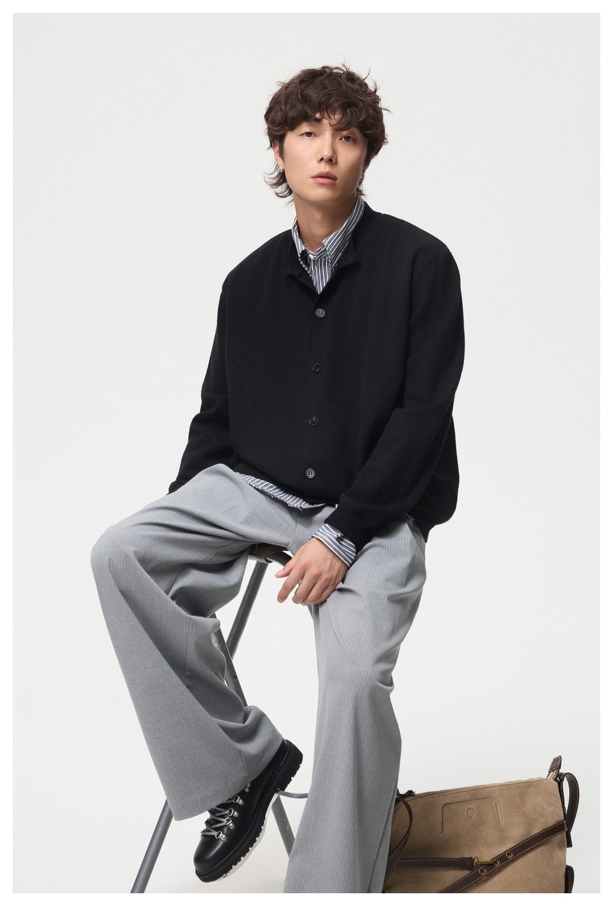 Seated model wearing a black button-front cardigan, striped shirt, and gray trousers.