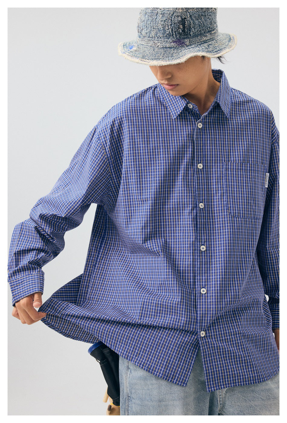 Model in an oversized blue check shirt with a bucket hat, pulling the hem with one hand