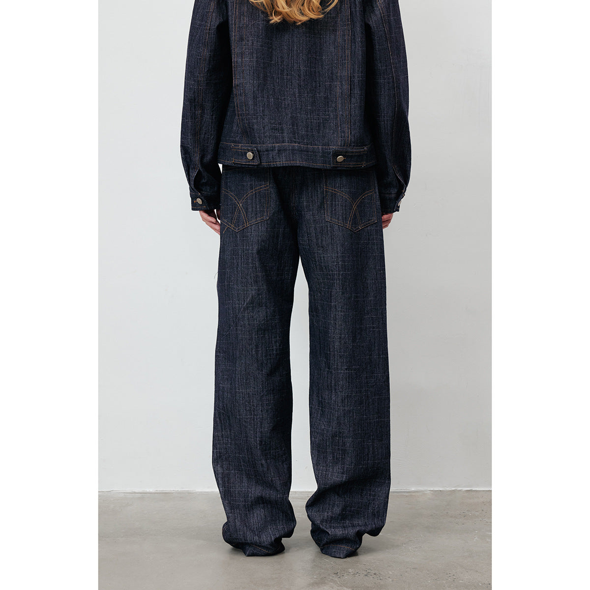 Rear view of a model wearing a dark indigo denim jacket and loose wide-leg jeans with curved-stitched back pockets.