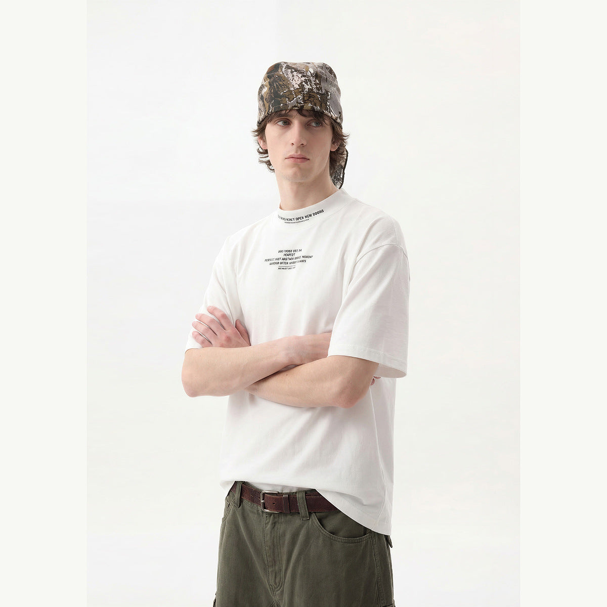 Model wearing a white oversized T-shirt with small black chest and collar text, olive trousers, and a camouflage headscarf.