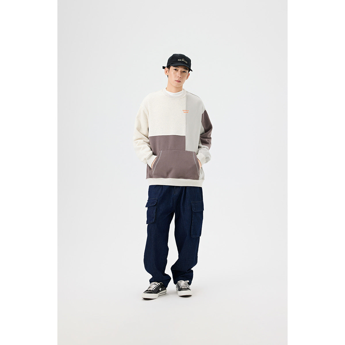 Full-length model wearing a color-block crewneck sweatshirt with a large front pocket and dark cargo jeans.
