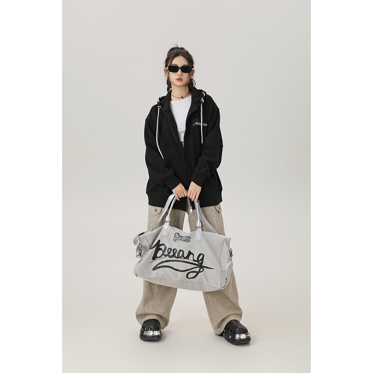 Model wearing an oversized black zip-up hoodie with white drawstrings, shown with loose beige cargo pants and a large gray