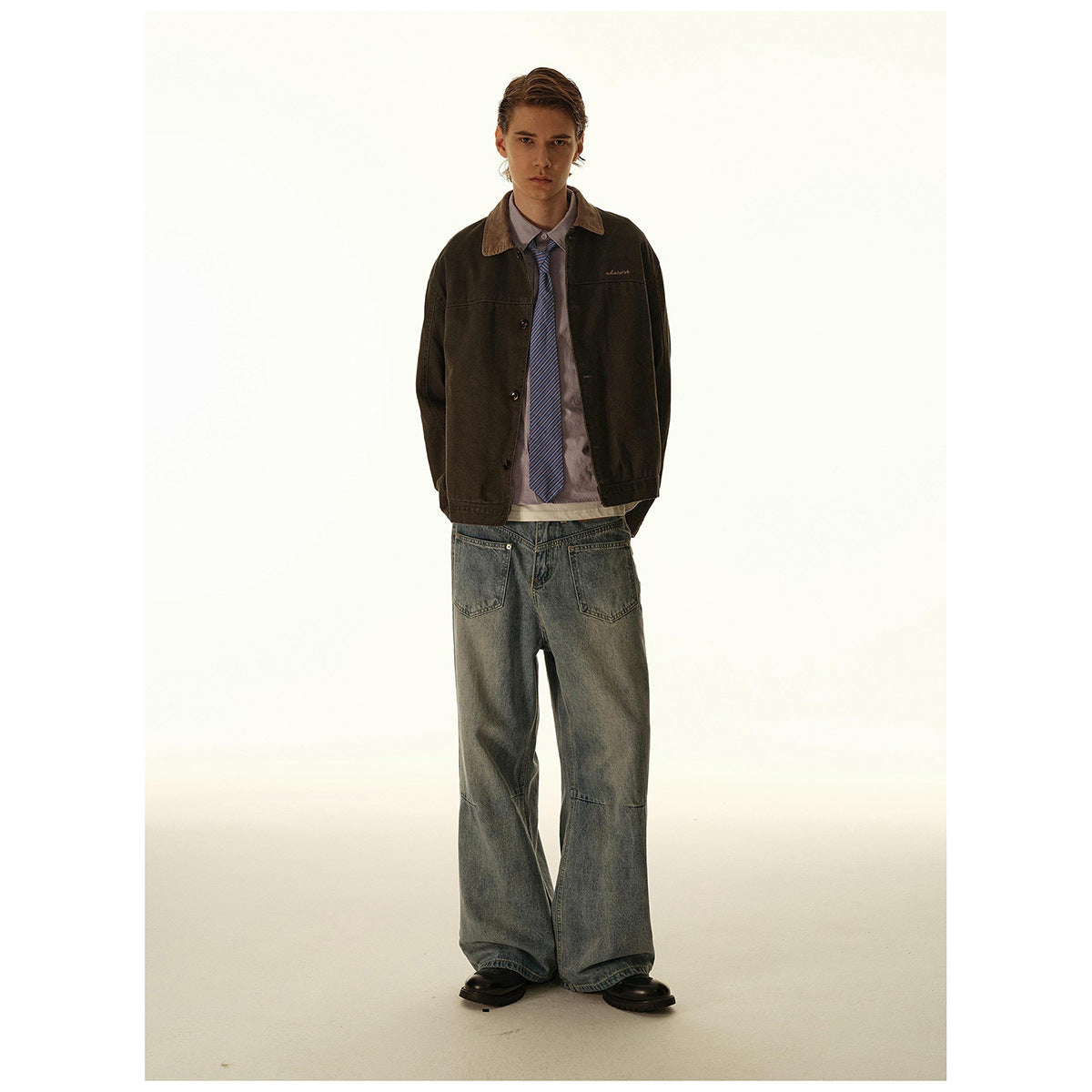 Model wearing a dark brown button-front jacket with a tan collar, striped tie, and loose faded jeans.