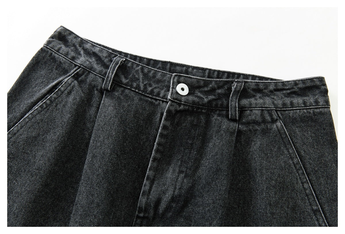 Angled close-up of the front waistband, button, belt loops and pleated front of black washed denim trousers.