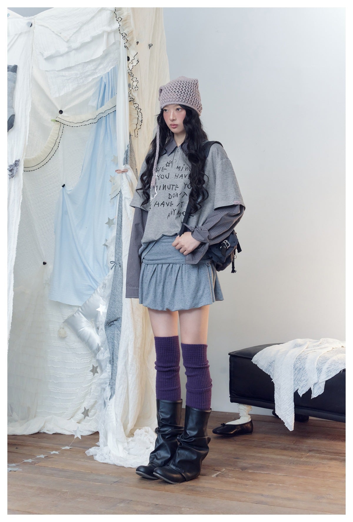 Full-length model wearing a gray printed layered top and mini skirt with a lavender hat and purple leg warmers.