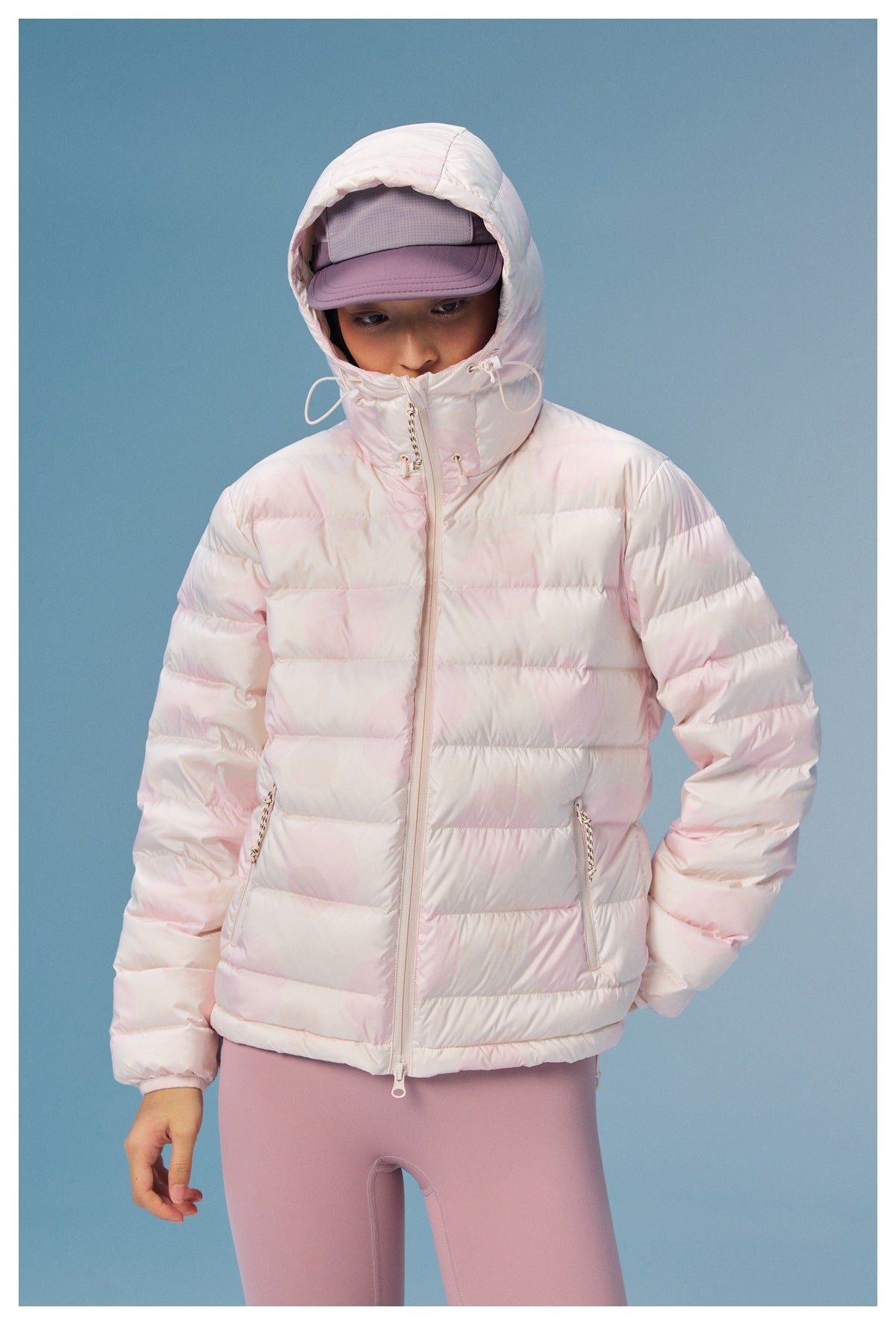 Model wearing a pale pink quilted hooded jacket with a high drawcord collar and zip pockets, paired with mauve leggings.