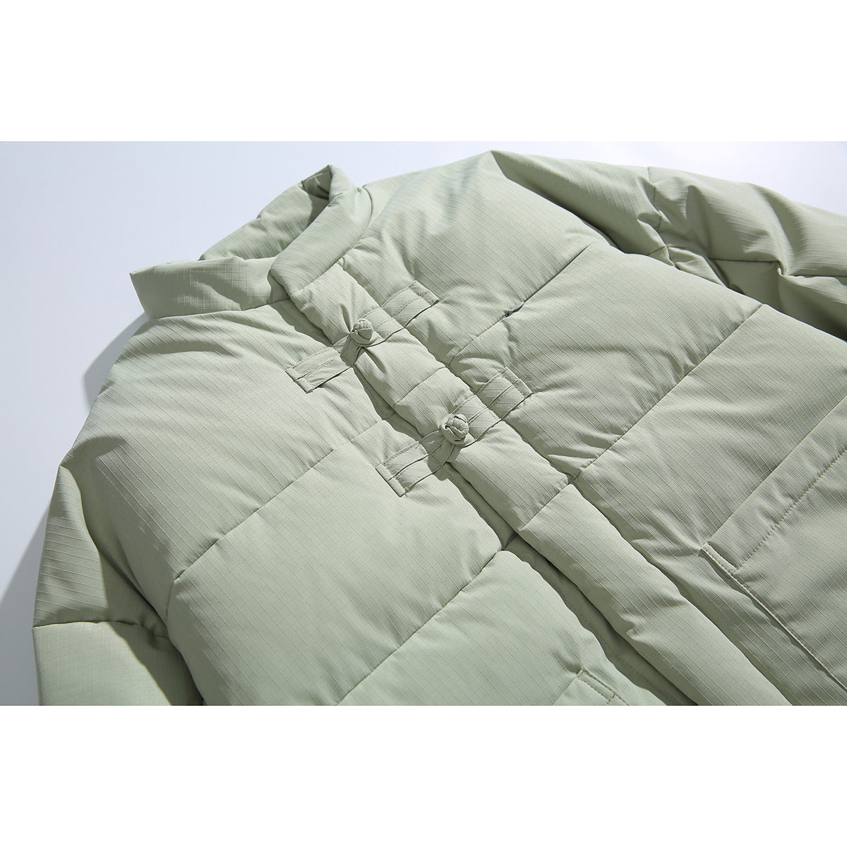 Close-up of a pale sage padded jacket with horizontal quilting, a high collar and fabric knot closures.