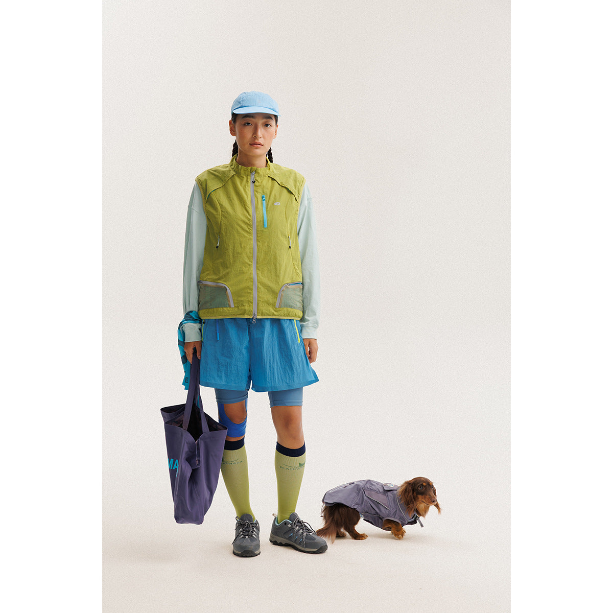 Model wearing a lime-green zip-front vest with blue trim, blue shorts and a pale long-sleeve top.