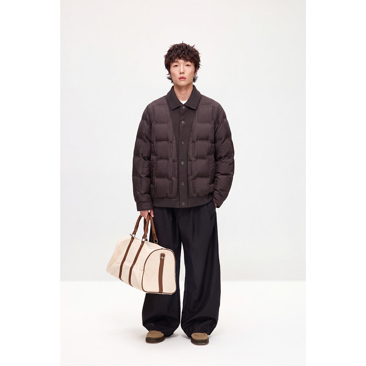 Full-length model wearing a dark brown quilted snap-front jacket with black trousers and carrying a cream duffel bag.