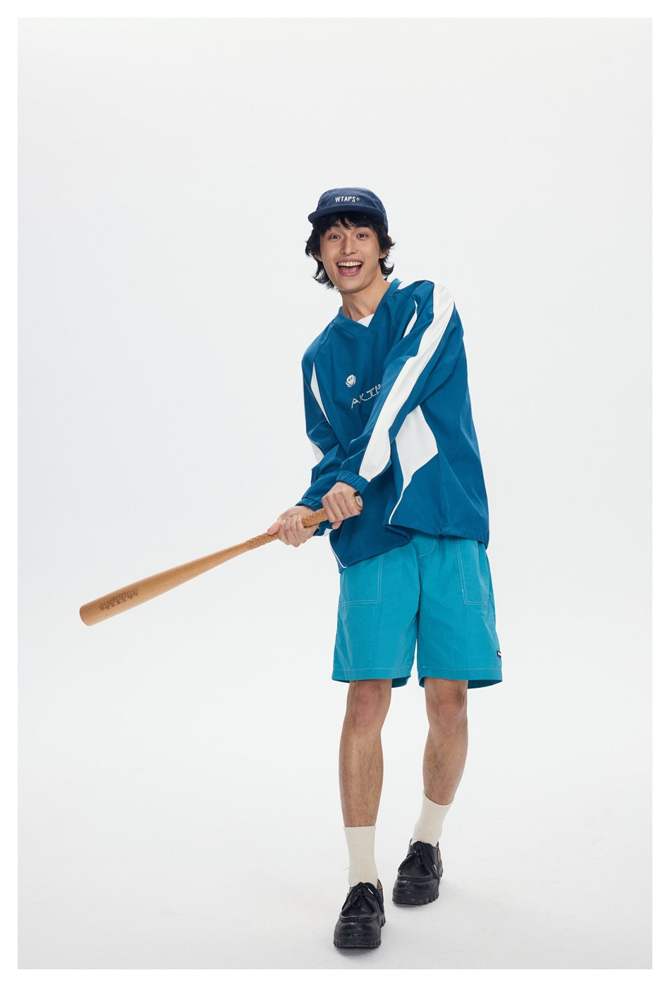 Full-length model wearing a teal-and-white pullover and teal shorts while holding a wooden bat.