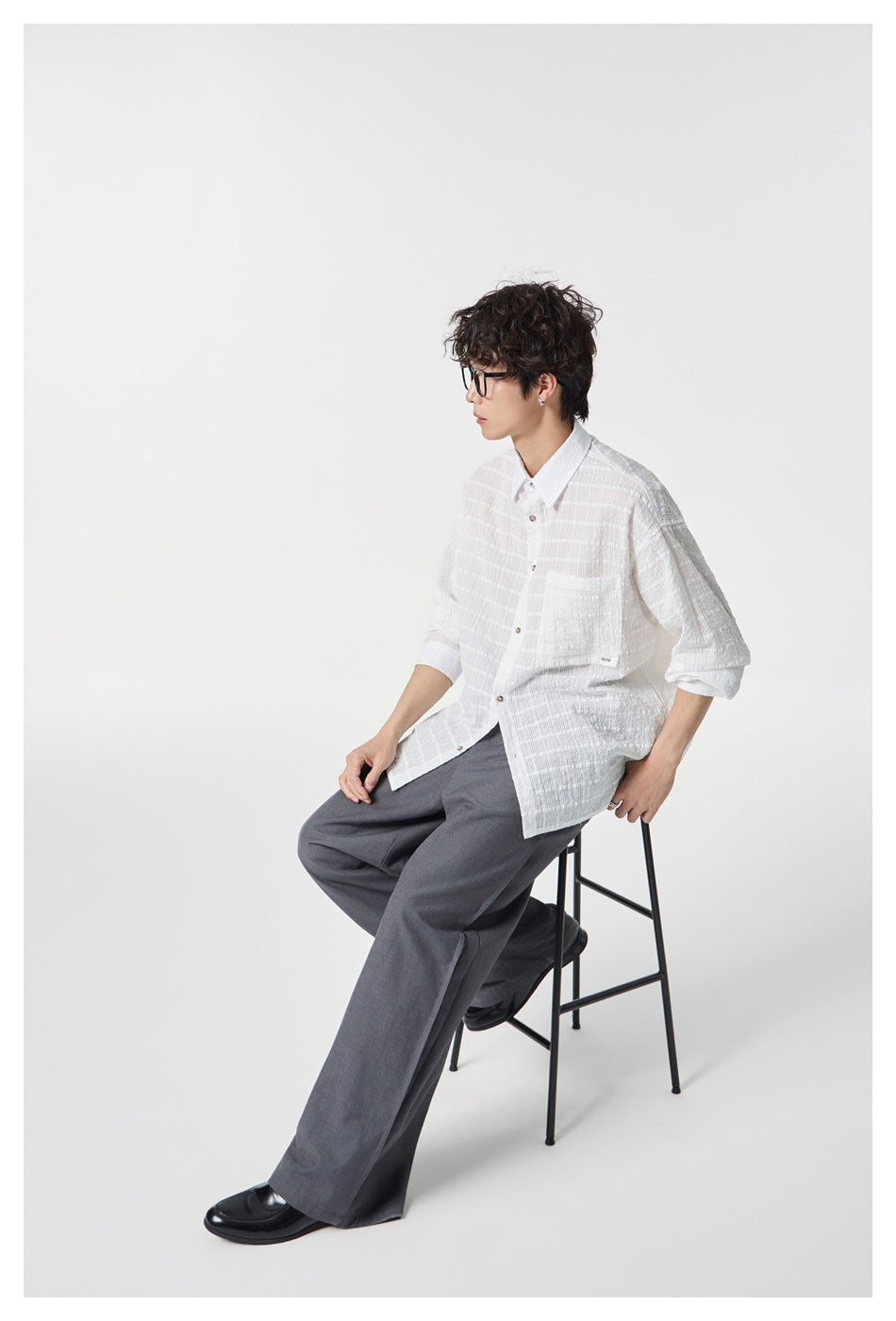 Seated model wearing a white textured button-up shirt with a chest pocket and gray wide-leg trousers.