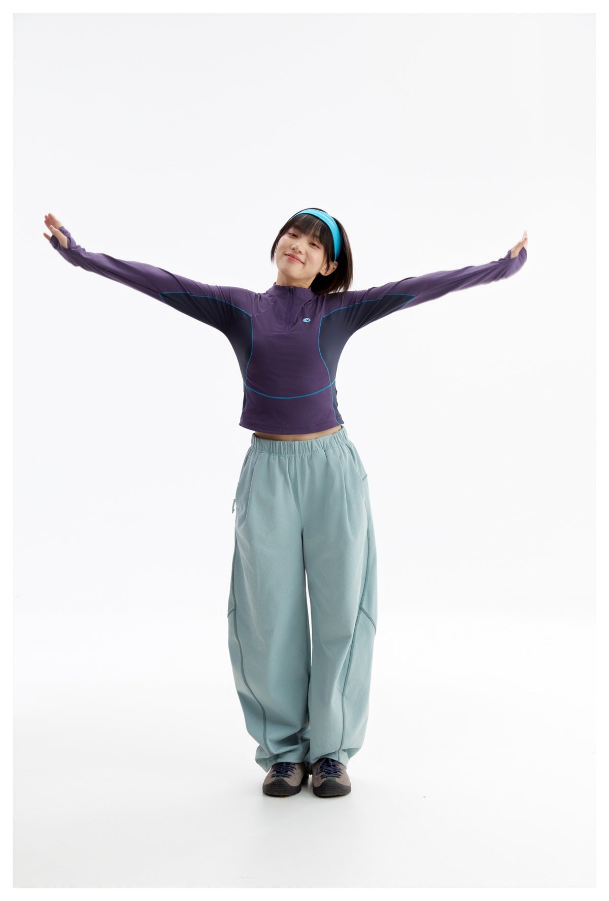Model wearing a purple long-sleeve quarter-zip top with teal piping and loose pale blue trousers.