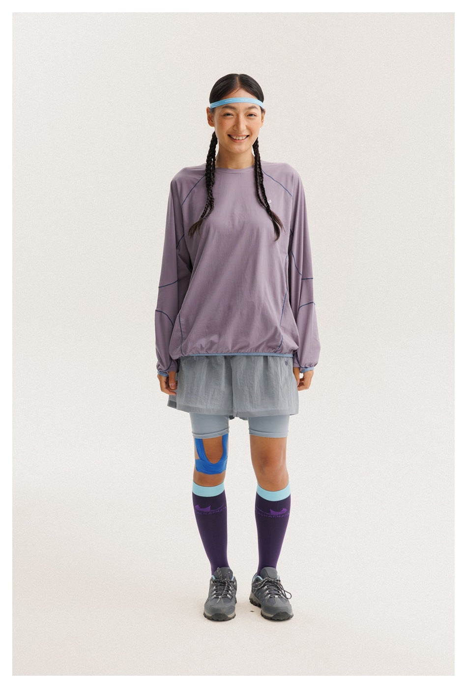 Front view of a woman wearing a loose lavender long-sleeve top with blue seam piping, gray running shorts, and purple knee
